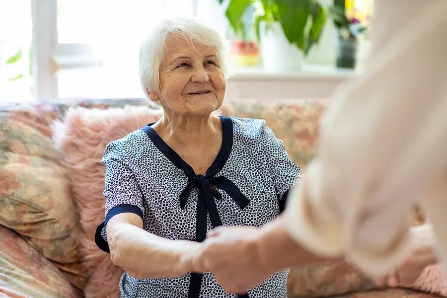 Elderly Lady Smiling Whilst A Carer Holds Her Hands, Helping Her To Stand