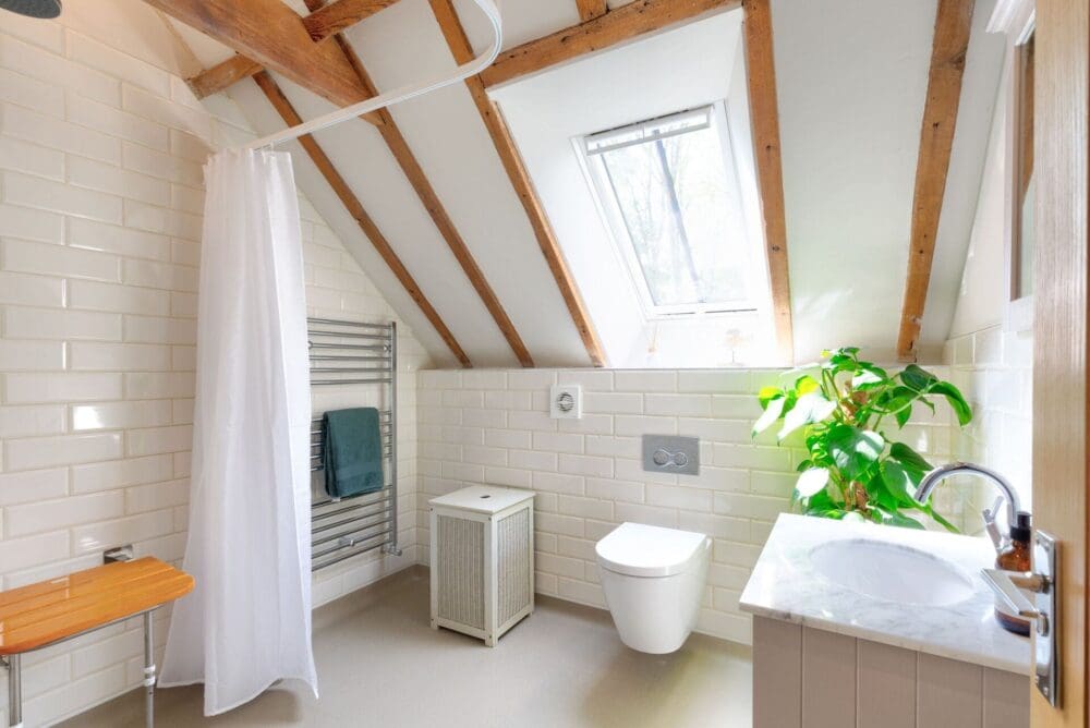 Mr And Mrs W's Bathroom After Renovation White, Clean Tiled Walls An Exposed Truss Slanted Roof, With Large Plant, Towel Rail, Toilet, Walk In Shower And Shower Seat