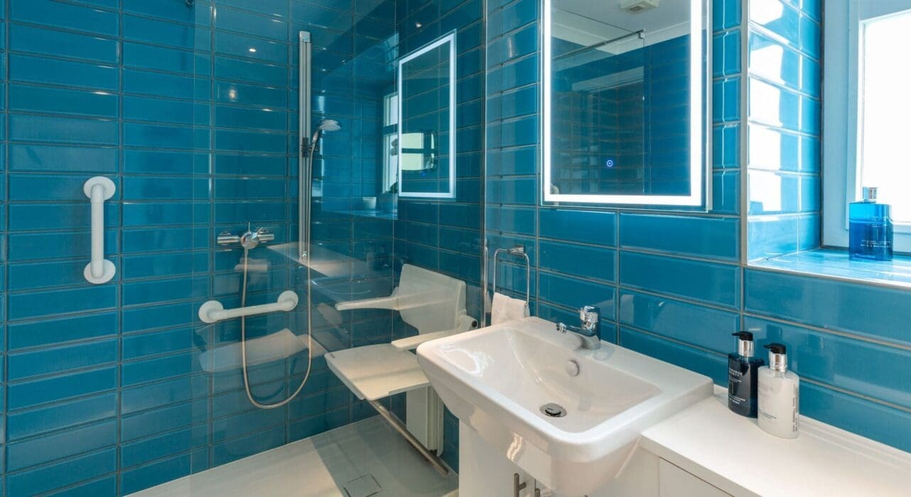 Mrs W's Bathroom After Renovation Glossy Blue Tiled Walls With Light Up Mirror, Contemporary White Sink And Cabinet, And Walk In Shower With Adjustable Shower Seat And Grab Rails (1)