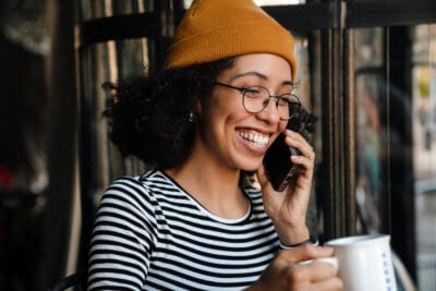 Young African American Woman On The Phone Smiling, Wearing A Beanie And Holding A Mug